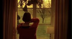 a woman standing in front of a window looking out at the grass and trees outside