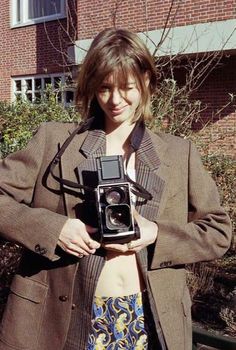 a woman holding an old camera in front of a brick building with bushes behind her