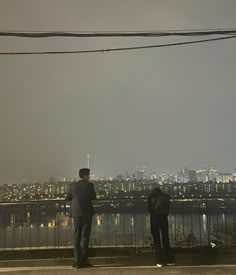 two people standing on the side of a road looking out over a city at night