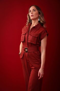 a woman standing in front of a red background wearing a red jumpsuit and earrings