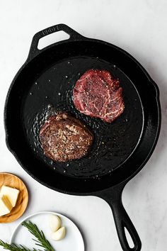 two steaks cooking in a cast iron skillet on a white countertop next to sliced cheese and herbs