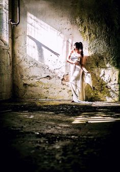 a woman in a white dress standing next to a brick wall with sunlight coming through the window