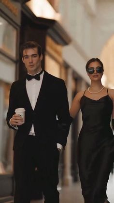 a man and woman in formal wear walking down the street with one holding a coffee cup