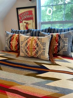 a bed topped with lots of pillows next to a window covered in blankets and throw pillows