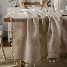 a dining room table covered with a beige linen blanket and tasseled chairs in the background