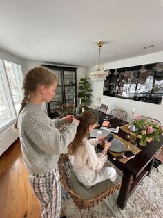 a woman sitting at a table cutting another woman's hair in front of a window