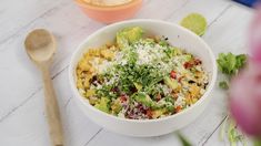 a white bowl filled with corn and vegetables next to a wooden spoon on top of a table