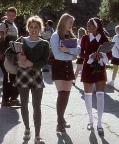 three girls in school uniforms are walking down the street with other people behind them and one girl is holding a book