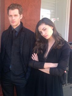 a man and woman standing next to each other in front of a building with their arms crossed