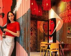 a woman standing next to a wall covered in red and black artwork with colorful lights hanging from it's sides