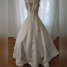 a wedding dress on display in front of a window with white curtains and wooden flooring