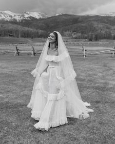 a woman wearing a wedding dress and veil standing in a field with mountains in the background