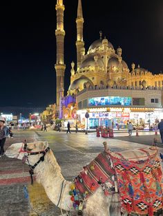 two camels are standing in front of an ornate building at night with people walking around