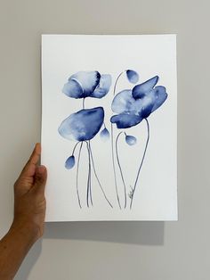 a hand holding up a piece of paper with watercolor flowers on it, in front of a white wall