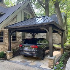 Elegant carport featuring a dark gray metal roof, beautifully complemented by tan stone accents and limestone piers. This open-sided structure shelters a black Volkswagen Tiguan, set against an old cottage-style house with black windows. The landscaping boasts lush greenery, colorful flowers, and two large decorative brown blocks enhancing the aesthetic. Carport Beside House, Beautiful Carport Ideas, Carport Driveway Ideas, Pergola As Carport, Cottage Carport Ideas, Adding A Carport To Front Of House, Carport With Privacy Wall