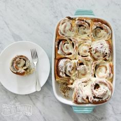 a blue dish filled with cinnamon rolls next to a white plate and fork on a marble surface