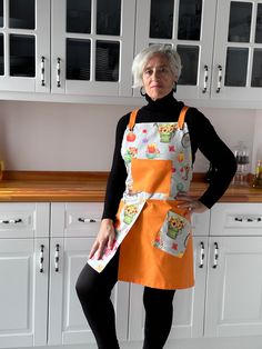 an older woman standing in the kitchen with her hands on her hips, wearing an orange apron and black leggings