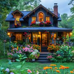 a blue house with lots of flowers in the front yard and lights on it's windows