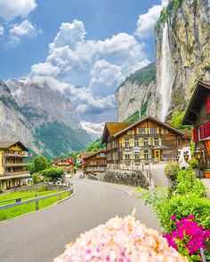 the road is lined with wooden buildings and flowers in front of some tall mountain peaks