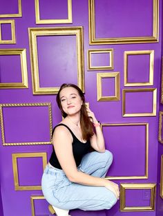 a woman sitting on top of a stool in front of a purple wall with gold frames