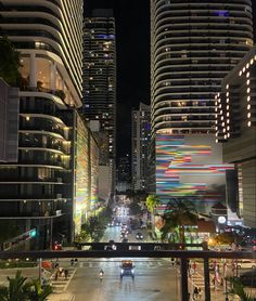 a city street at night with tall buildings in the background and cars driving on it