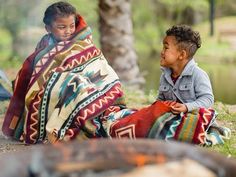 two children wrapped in blankets sitting on the ground next to a fire pit and trees