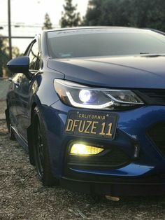 the front end of a blue car with its lights turned on and license plate showing