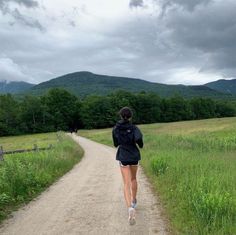 a woman running down a dirt road in the middle of a field with mountains behind her