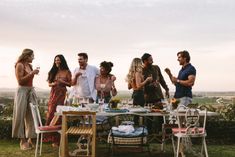 a group of people standing around a table with food and drinks on it in the grass