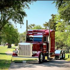 a large red semi truck driving down a street next to a lush green park on a sunny day