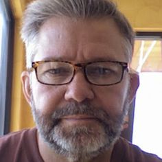 a man with glasses and a beard is looking at the camera while standing in front of a window