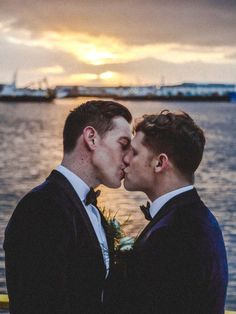 two men in tuxedos are kissing each other by the water at sunset or sunrise