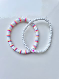 two white bracelets with multicolored beads and a heart on the clasp are sitting next to each other