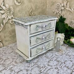 a white dresser with three drawers sitting on top of a lace covered tablecloth next to flowers