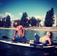 two shirtless men on a paddle boat in the water with buildings in the background