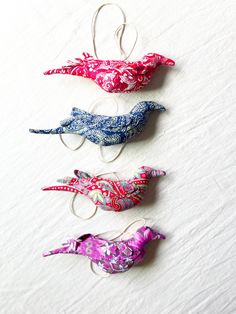 three small birds are hanging from hooks on a white tablecloth covered surface, one is pink, the other is blue