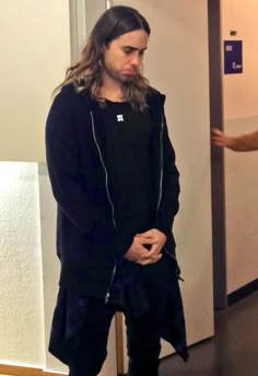 a man with long hair standing in front of an open door and wearing black pants