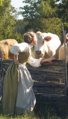 a woman in an old fashioned dress petting a cow on the nose while standing next to a fence