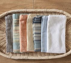 six towels folded in different colors on a wicker basket with wood flooring behind them