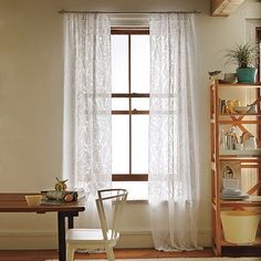 a room with a table, chair and window covered in white lace curtaines on the windowsill