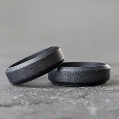 two black wedding bands sitting next to each other on top of a stone floor with grey background