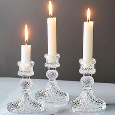 three crystal candlesticks with one lit and the other turned on, in front of a gray background