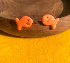 two small orange fish shaped earrings sitting on top of a wooden bowl next to a yellow cloth