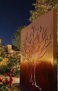 a large metal sculpture with a tree on it's side in front of flowers