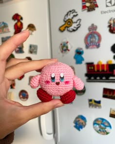 a hand holding a small pink crocheted animal in front of a refrigerator with magnets on it