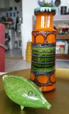 an orange and green vase sitting on top of a table next to a green fish figurine