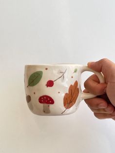 a hand holding a coffee cup decorated with leaves and mushrooms on white background, close up