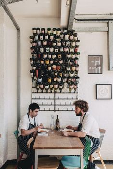 two men sitting at a table in front of a wall with wine bottles on it