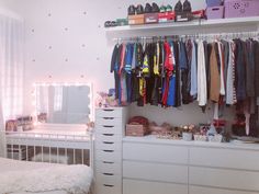 a white dresser sitting next to a bed in a bedroom under a window with lots of clothes hanging on it