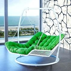 a white swing chair with green cushions in front of a large window overlooking the ocean